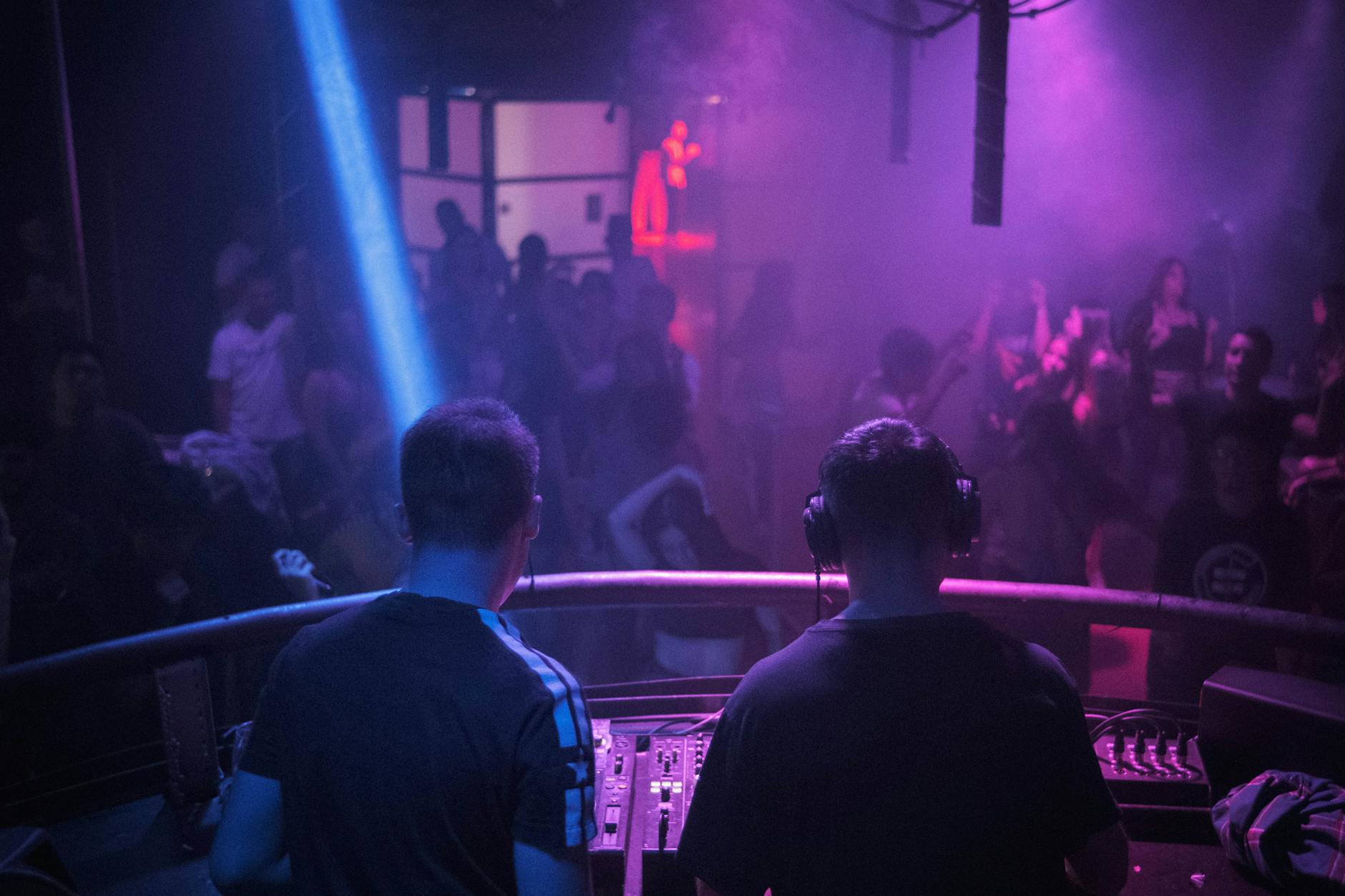 A lively night club scene with a DJ mixing music amidst vibrant lights and dancing crowd.