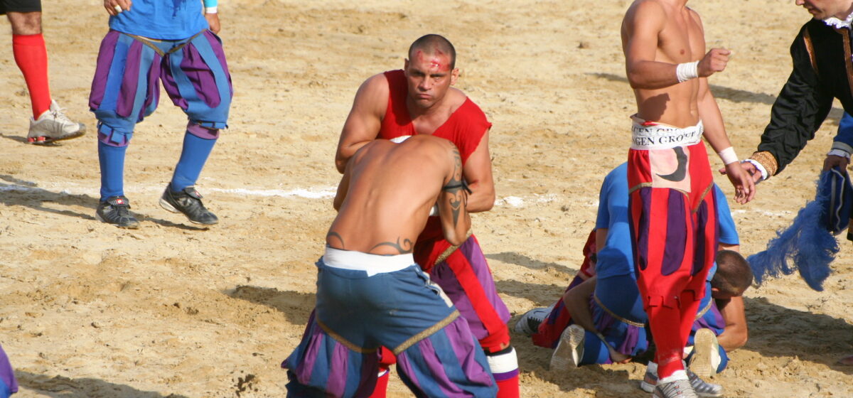 Calcio Storico players in blue and red Renaissance breeches grappling on the sand of Piazza Santa Croce, one with a bloodied forehead, an official in costume at the side