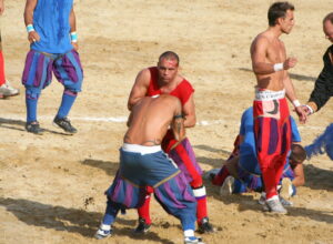 Calcio Storico players in blue and red Renaissance breeches grappling on the sand of Piazza Santa Croce, one with a bloodied forehead, an official in costume at the side