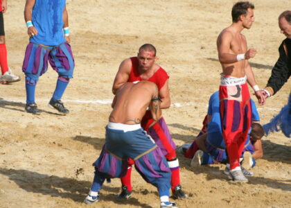 Calcio Storico players in blue and red Renaissance breeches grappling on the sand of Piazza Santa Croce, one with a bloodied forehead, an official in costume at the side
