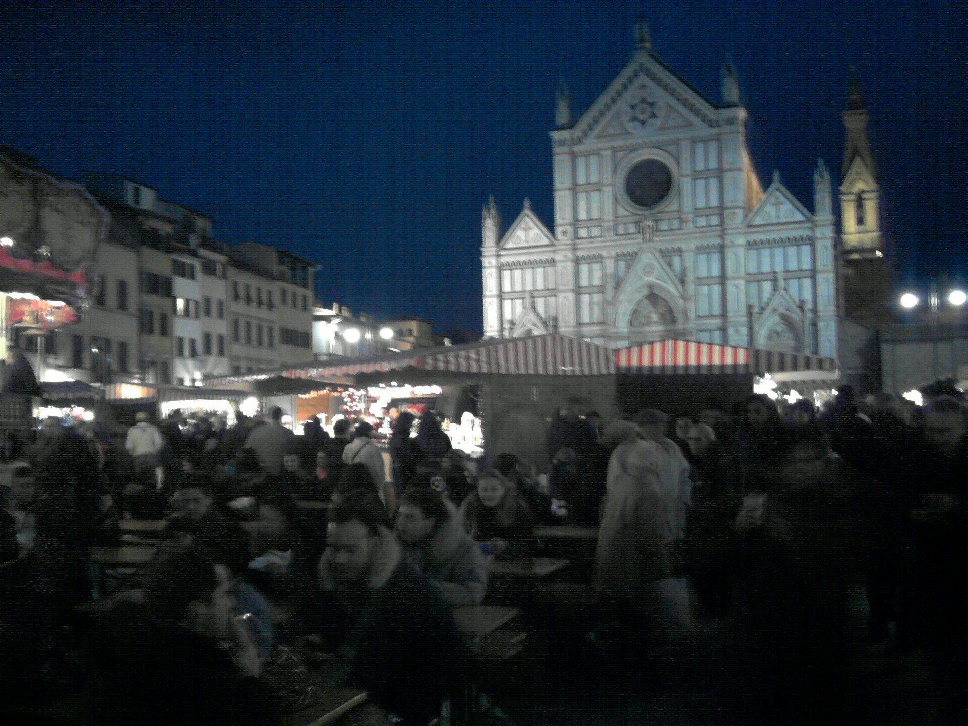 Wooden chalets with red and white striped awnings filling Piazza Santa Croce at night, crowds at trestle tables, the floodlit basilica facade behind