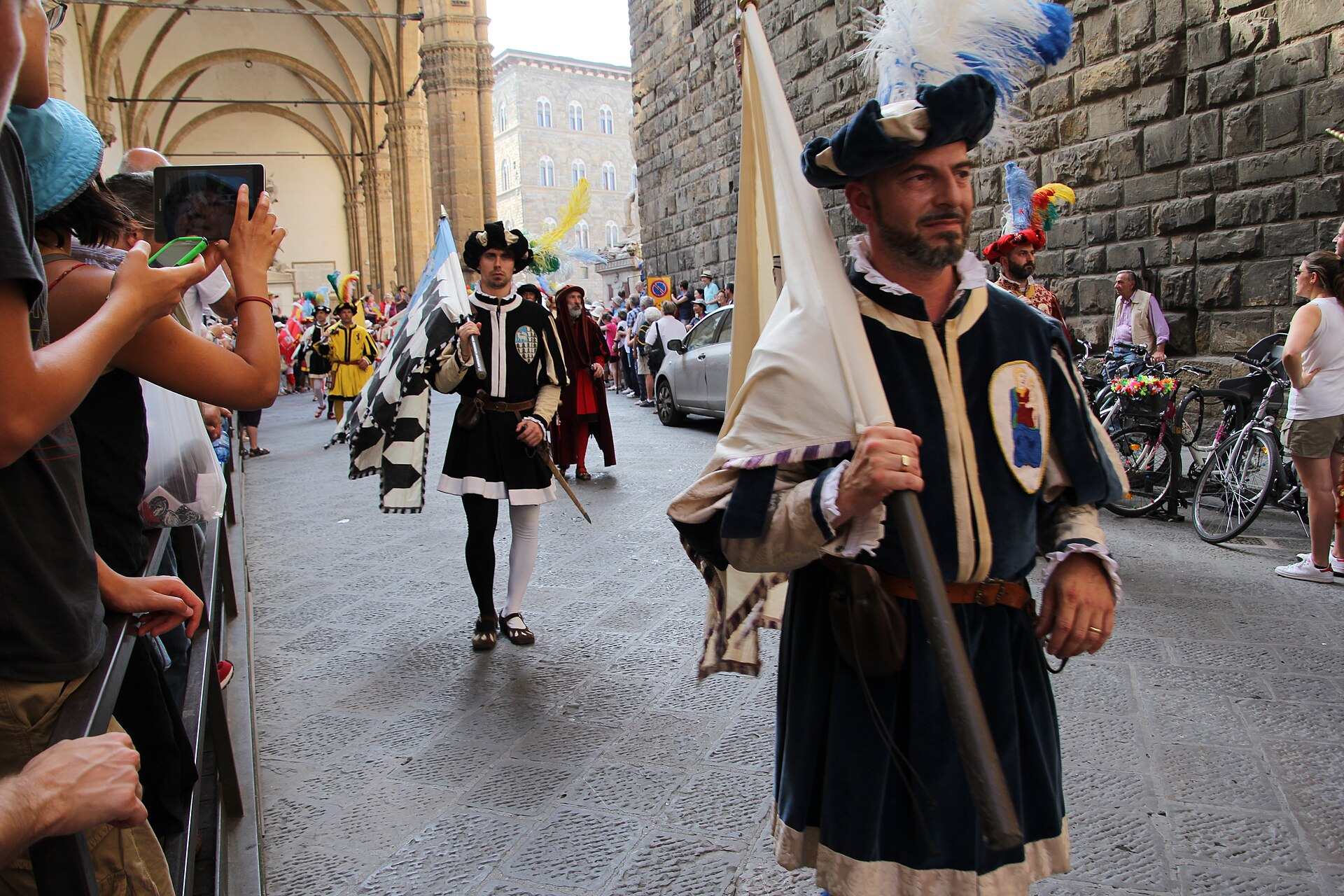 Flag-bearers of the Corteo Storico della Repubblica Fiorentina in Renaissance costume walking past a stone loggia while spectators film them