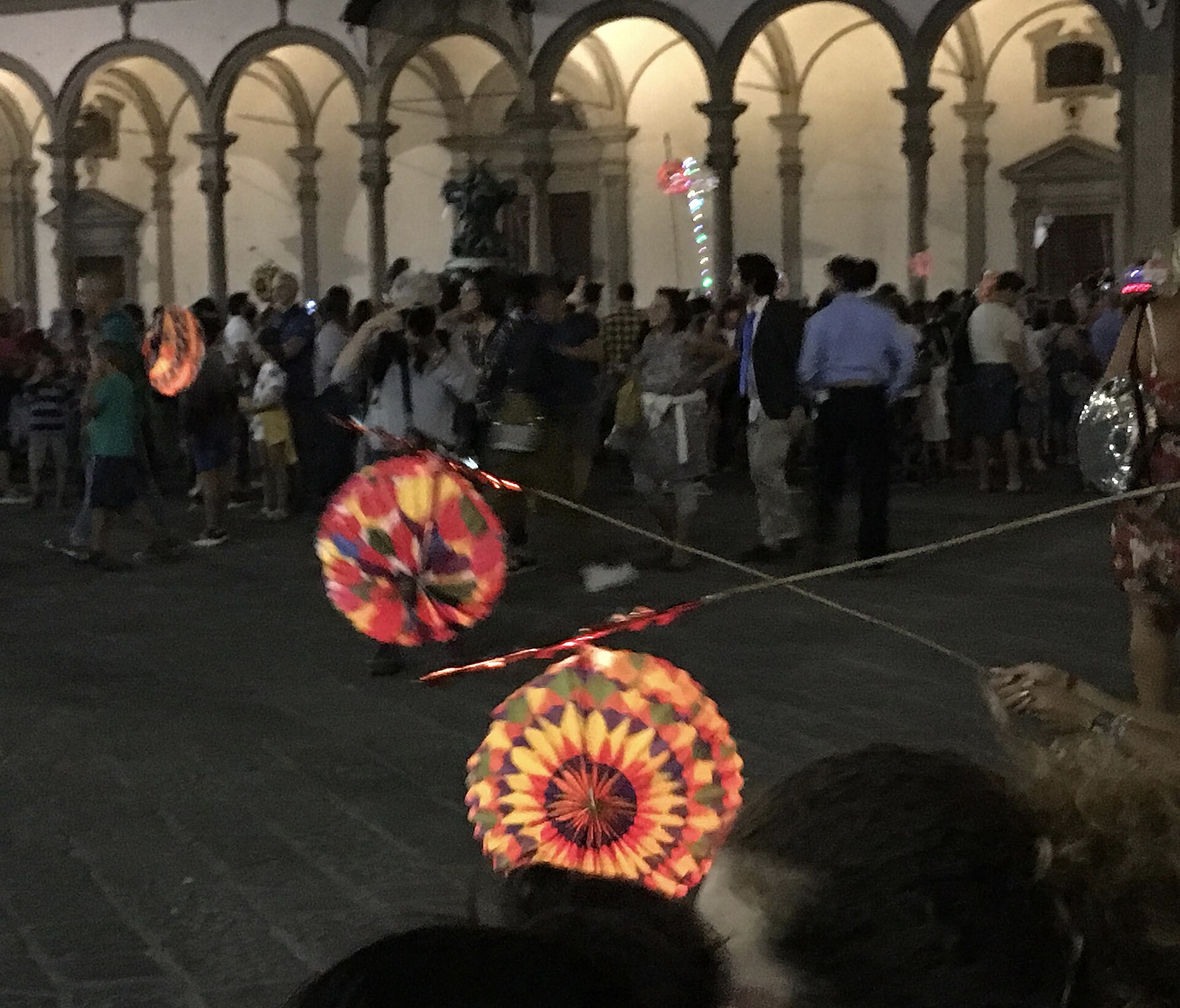 Brightly coloured paper lanterns on poles carried through a crowd at night under the arcades of Piazza Santissima Annunziata