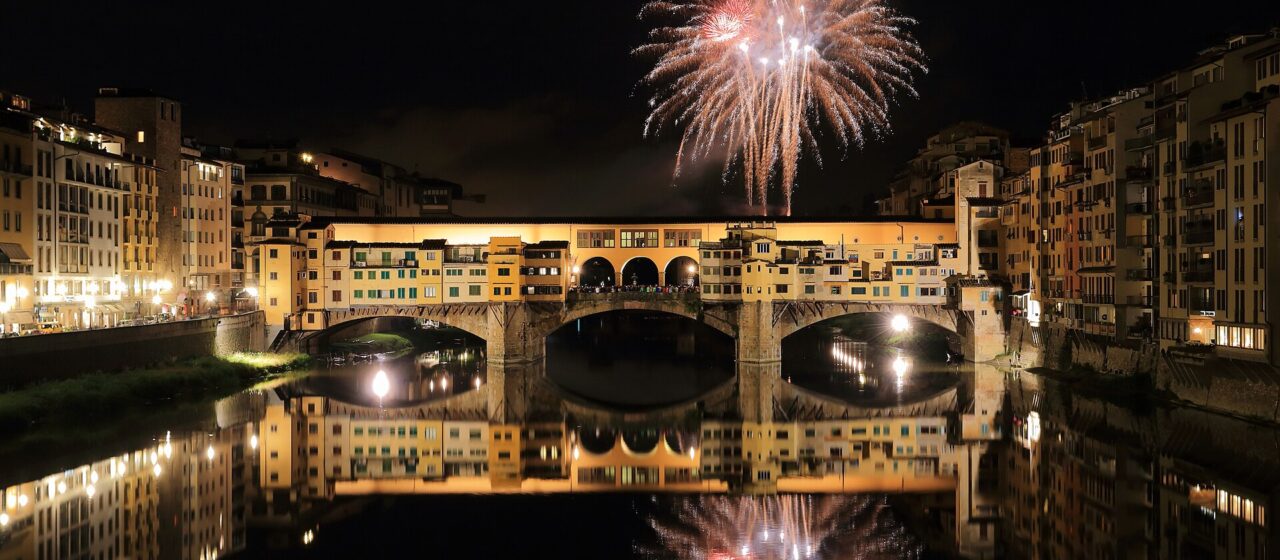A firework bursting in the night sky above Ponte Vecchio, the bridge lit gold and reflected in the black water of the Arno