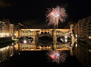 A firework bursting in the night sky above Ponte Vecchio, the bridge lit gold and reflected in the black water of the Arno