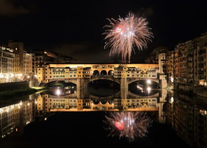 A firework bursting in the night sky above Ponte Vecchio, the bridge lit gold and reflected in the black water of the Arno