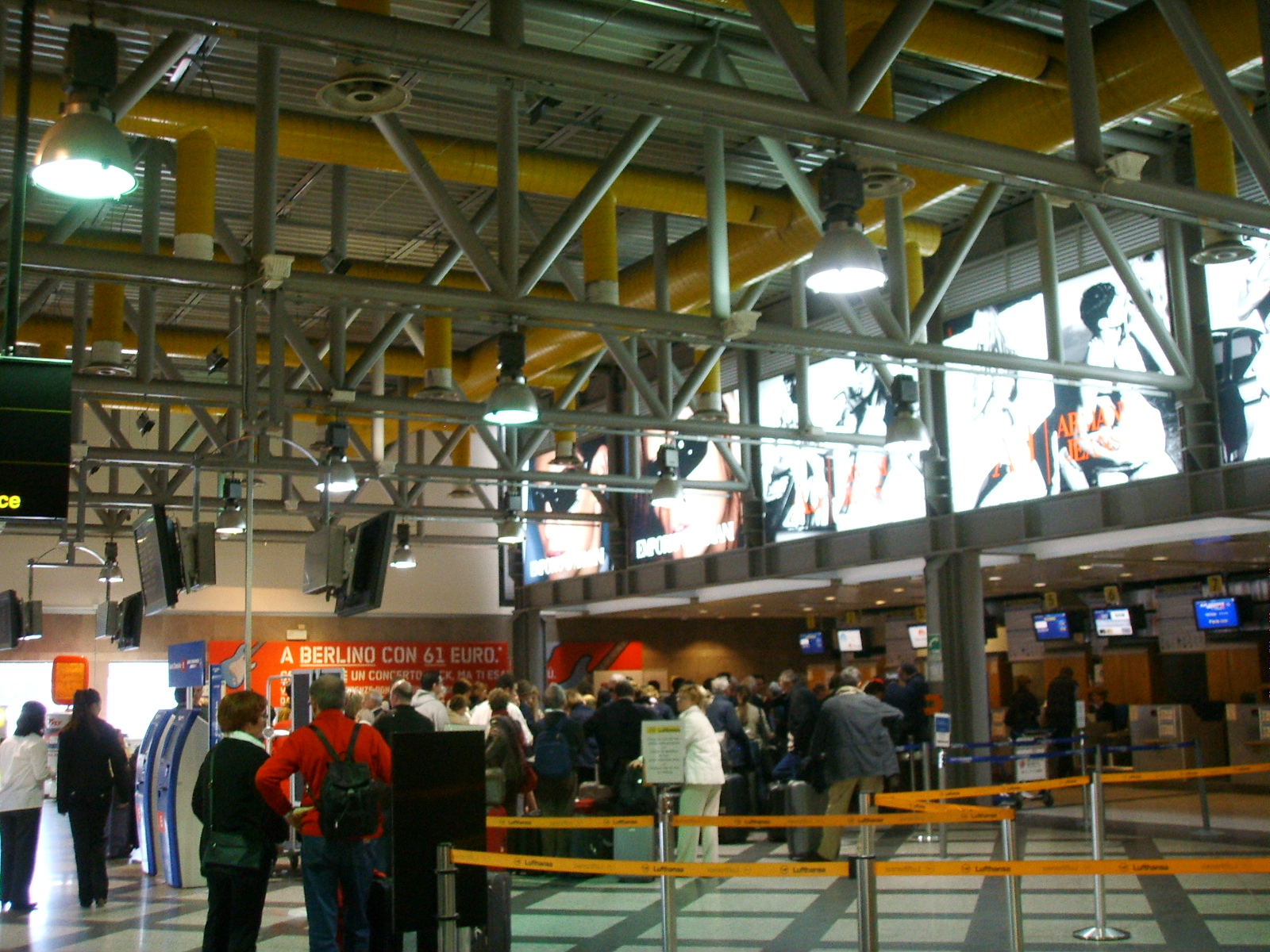The check-in hall at Florence airport with steel roof trusses, exposed yellow ducting, rows of check-in desks and passengers queueing behind barriers