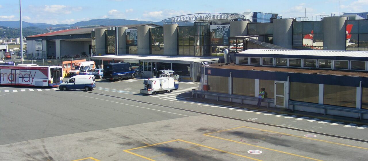 The main terminal building at Florence Amerigo Vespucci airport seen from the apron, with concrete buttresses, glass frontage, ground vehicles and the Tuscan hills behind
