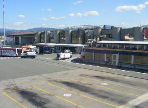 The main terminal building at Florence Amerigo Vespucci airport seen from the apron, with concrete buttresses, glass frontage, ground vehicles and the Tuscan hills behind