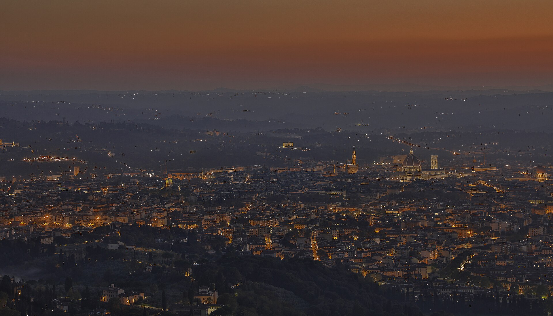 Florence at dusk seen from the hills, the Duomo and Palazzo Vecchio tower lit above a haze of orange street lights under an orange sky