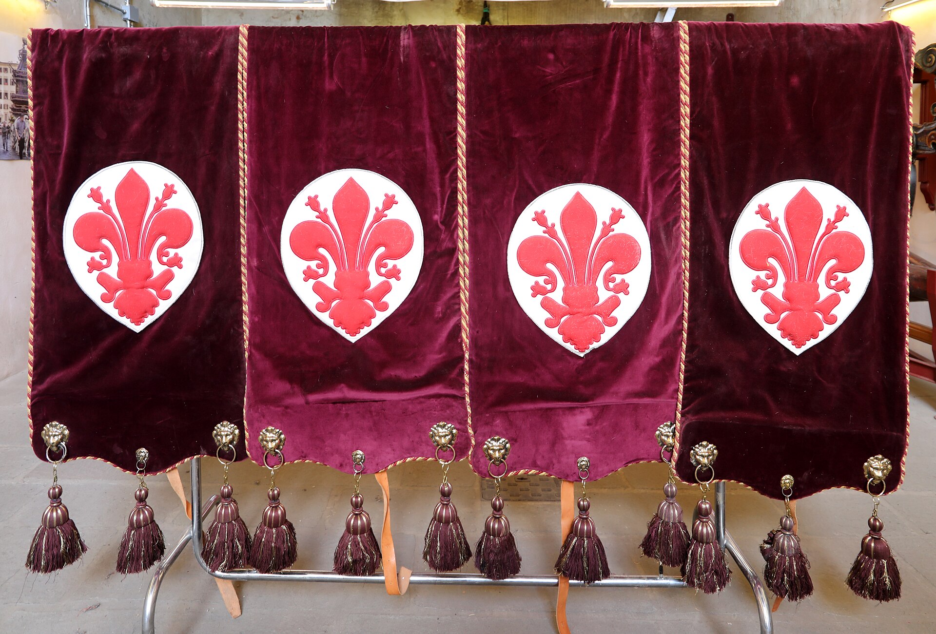 Four deep red velvet cloths hanging on a rack, each embroidered with the red fleur-de-lis of Florence on a white ground, finished with tassels and lion-head fittings