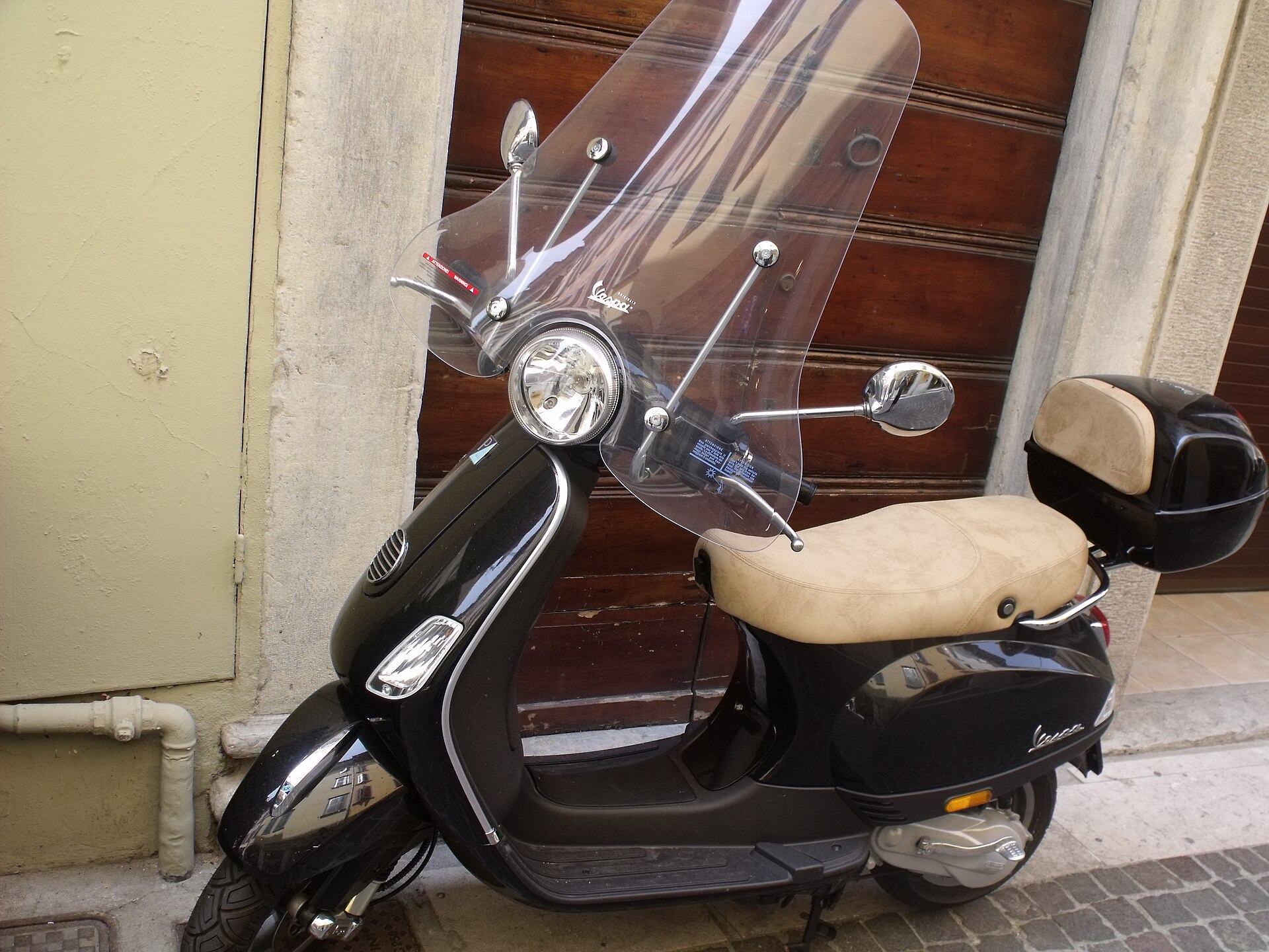 A black modern Piaggio Vespa with a clear windscreen and top box parked against a wooden door in an Italian street