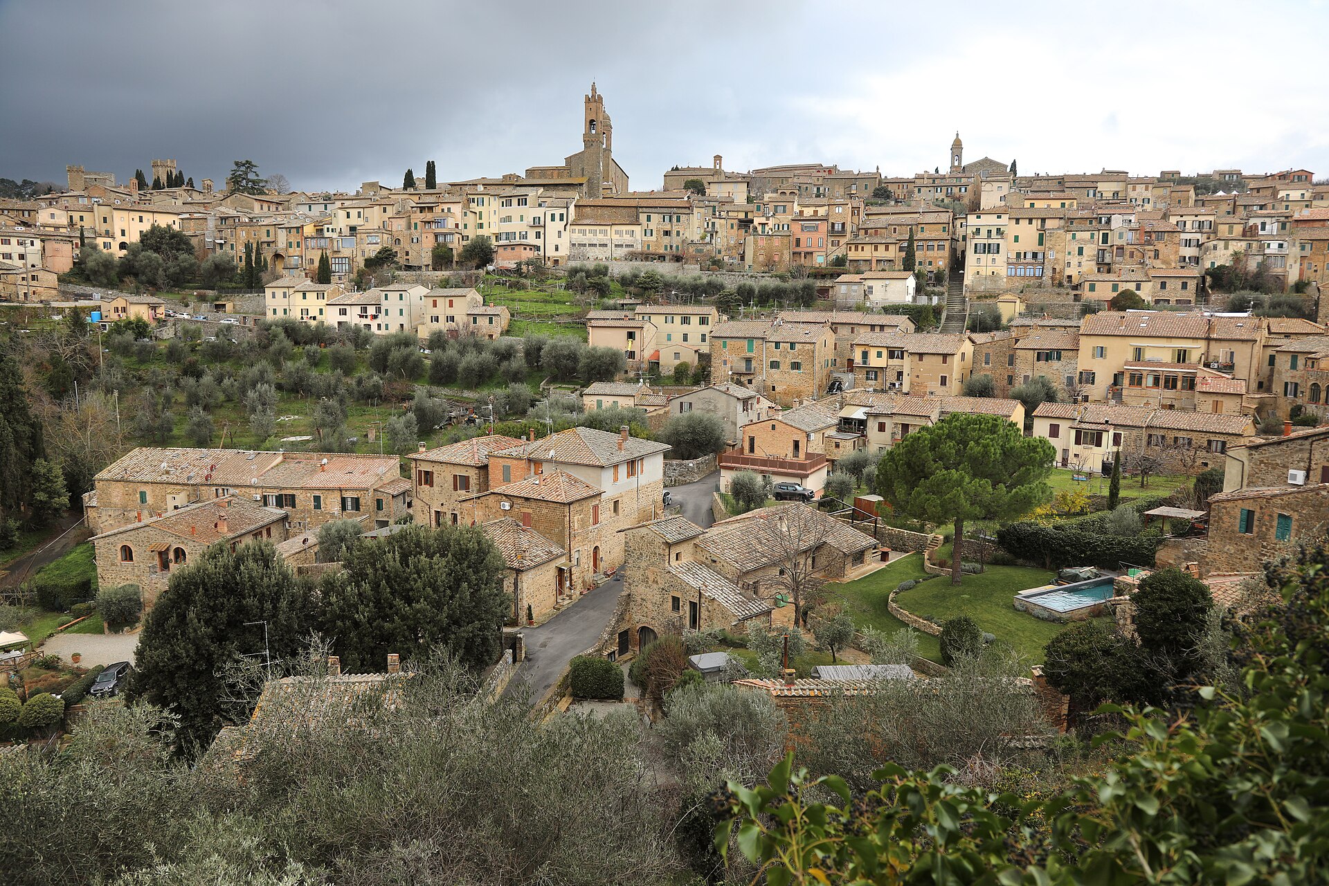 The hill town of Montalcino under a dark storm sky, stone houses stacked up the slope with olive terraces and cypresses below