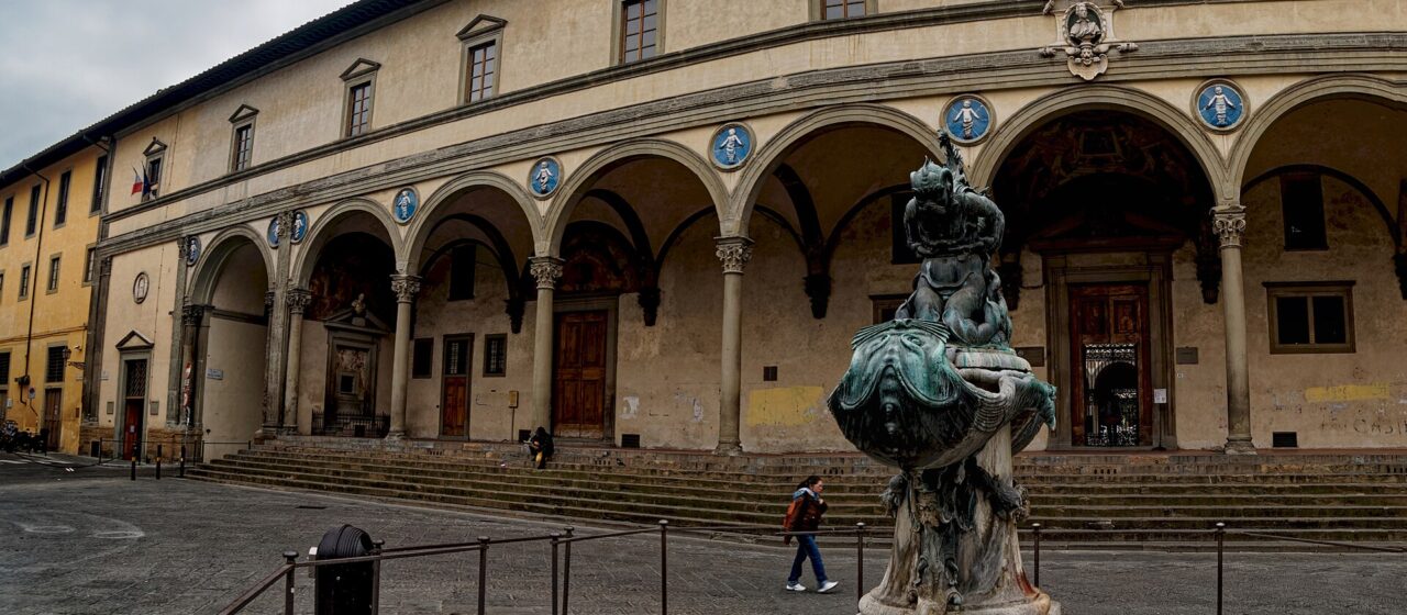Piazza della Santissima Annunziata under a grey sky, the arcade of the Ospedale degli Innocenti with its blue della Robbia roundels, a bronze sea-monster fountain in the foreground and one person walking across an otherwise empty square