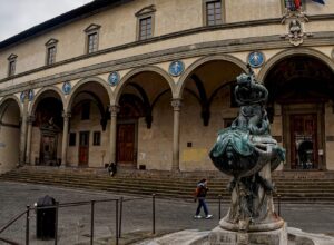 Piazza della Santissima Annunziata under a grey sky, the arcade of the Ospedale degli Innocenti with its blue della Robbia roundels, a bronze sea-monster fountain in the foreground and one person walking across an otherwise empty square