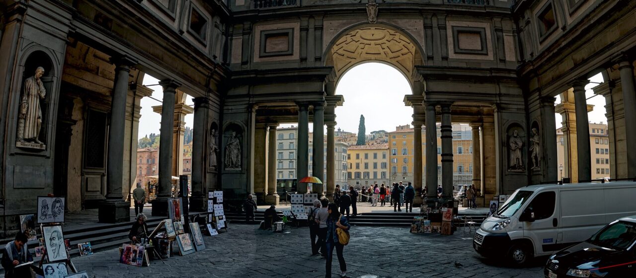 The long colonnaded courtyard of the Uffizi looking towards the arch that frames the Arno, with street artists and their easels along one side