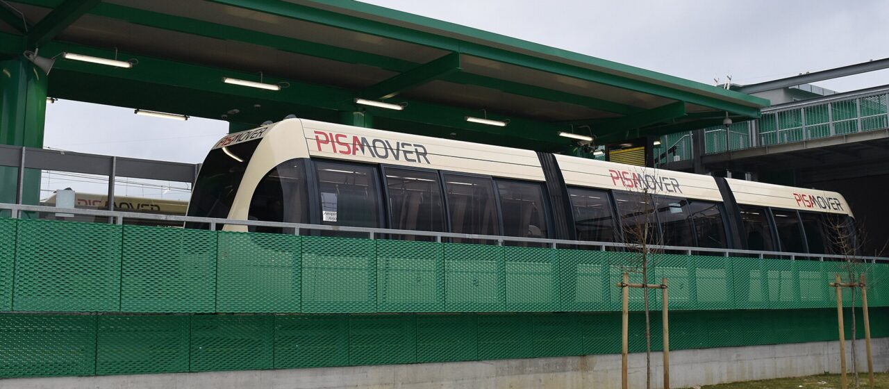 A cream and red PisaMover automated shuttle vehicle standing at its station platform under a green steel canopy