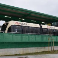 A cream and red PisaMover automated shuttle vehicle standing at its station platform under a green steel canopy