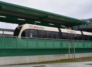 A cream and red PisaMover automated shuttle vehicle standing at its station platform under a green steel canopy