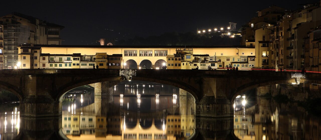 Ponte Santa Trinita in the foreground and Ponte Vecchio behind it at night, both lit and reflected in the still water of the Arno