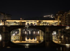 Ponte Santa Trinita in the foreground and Ponte Vecchio behind it at night, both lit and reflected in the still water of the Arno