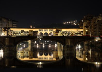 Ponte Santa Trinita in the foreground and Ponte Vecchio behind it at night, both lit and reflected in the still water of the Arno