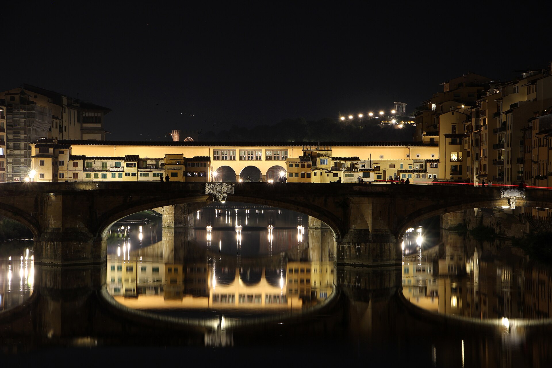 Ponte Santa Trinita in the foreground and Ponte Vecchio behind it at night, both lit and reflected in the still water of the Arno