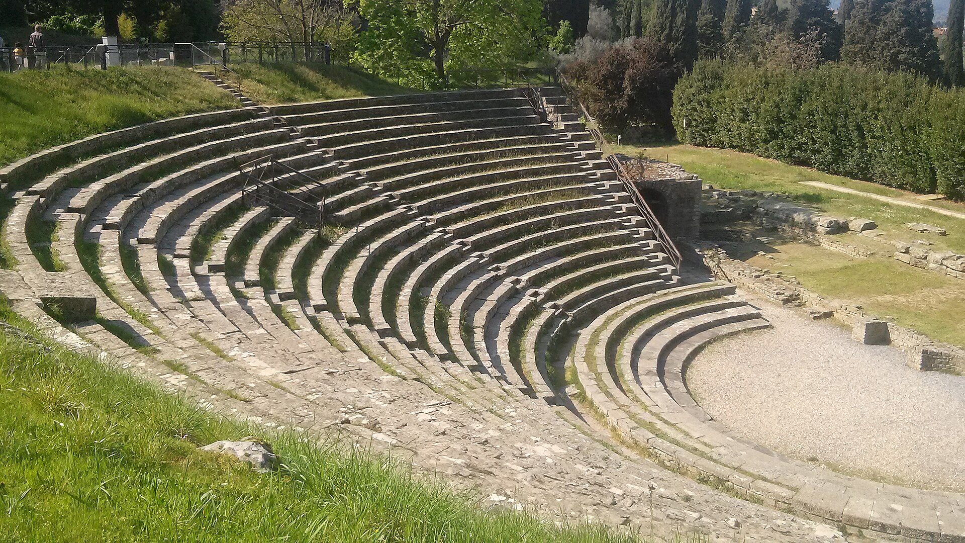 The curved stone tiers of the Roman theatre at Fiesole under sunlight, with cypresses and hedges behind and the excavated stage area at the bottom