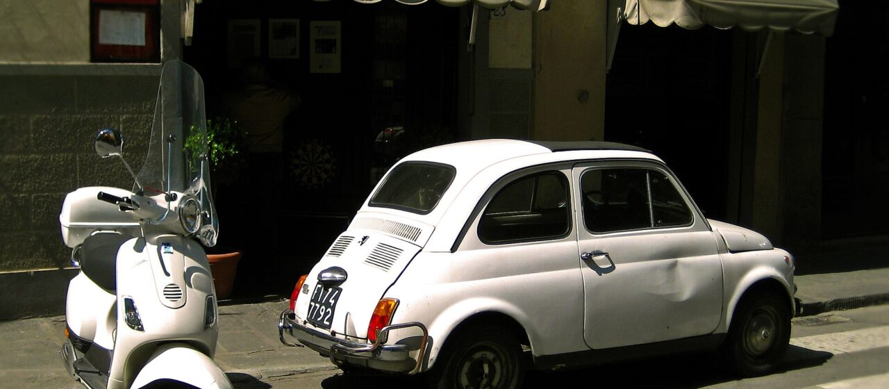 A white Vespa parked beside a vintage white Fiat 500 outside a trattoria in a Florentine street