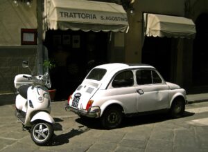 A white Vespa parked beside a vintage white Fiat 500 outside a trattoria in a Florentine street