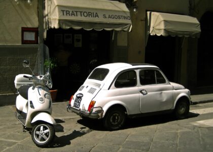 A white Vespa parked beside a vintage white Fiat 500 outside a trattoria in a Florentine street