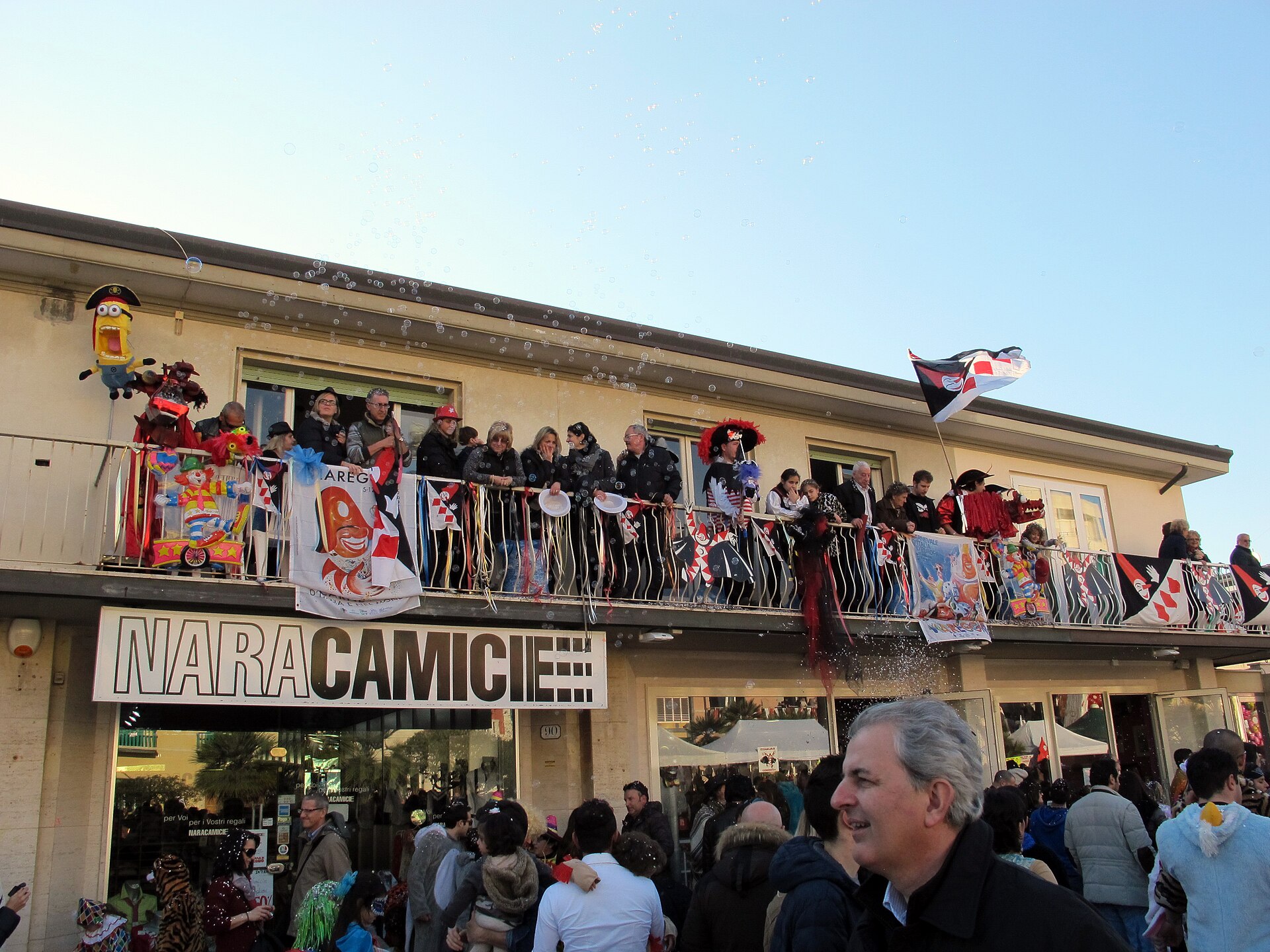 A carnival crowd on the Viareggio seafront, with people in costume on a decorated balcony above a shop, soap bubbles in the air
