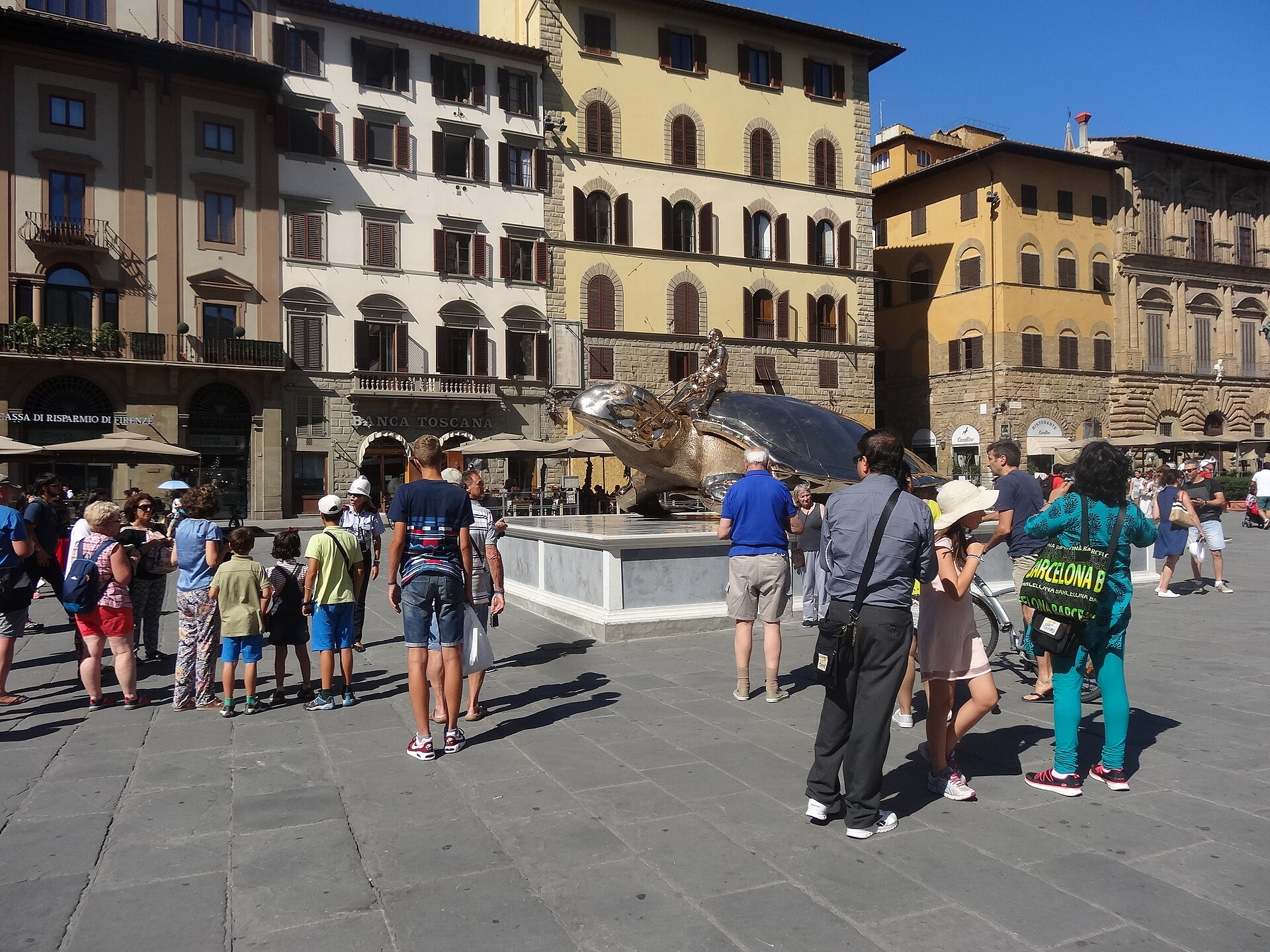 Adults and children gather around a large turtle sculpture on Florence’s Piazza della Signoria beneath sunlit palazzo facades.