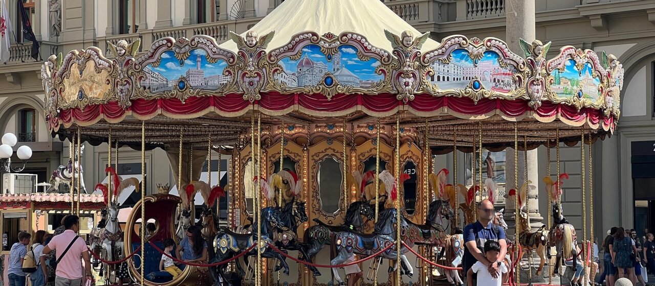 Adults and children gather around the painted-horse carousel in Florence’s Piazza della Repubblica beneath its ornate canopy.