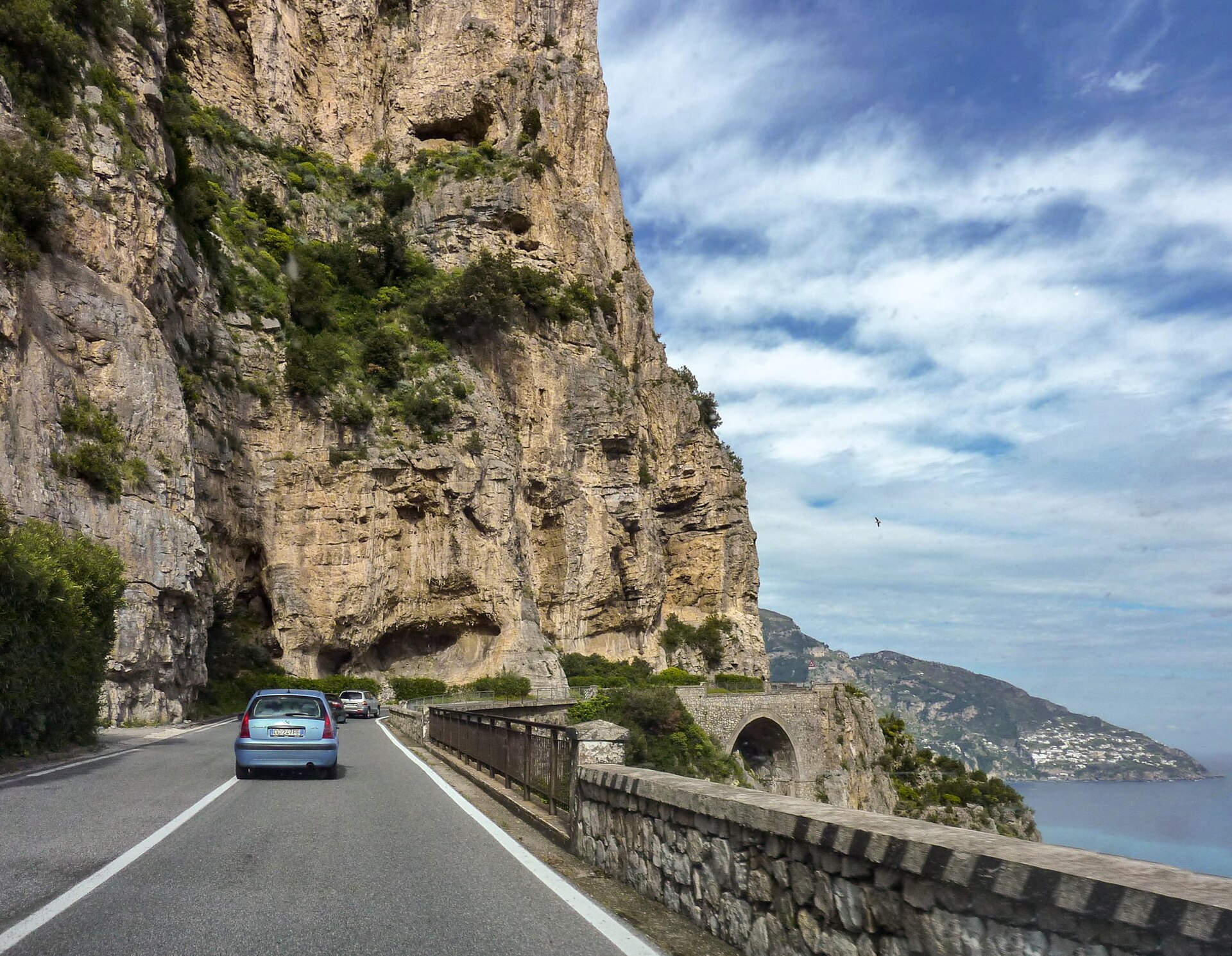 The Amalfi Coast road cutting under an overhanging limestone cliff, with a stone parapet on the seaward side and cars driving away along the narrow carriageway