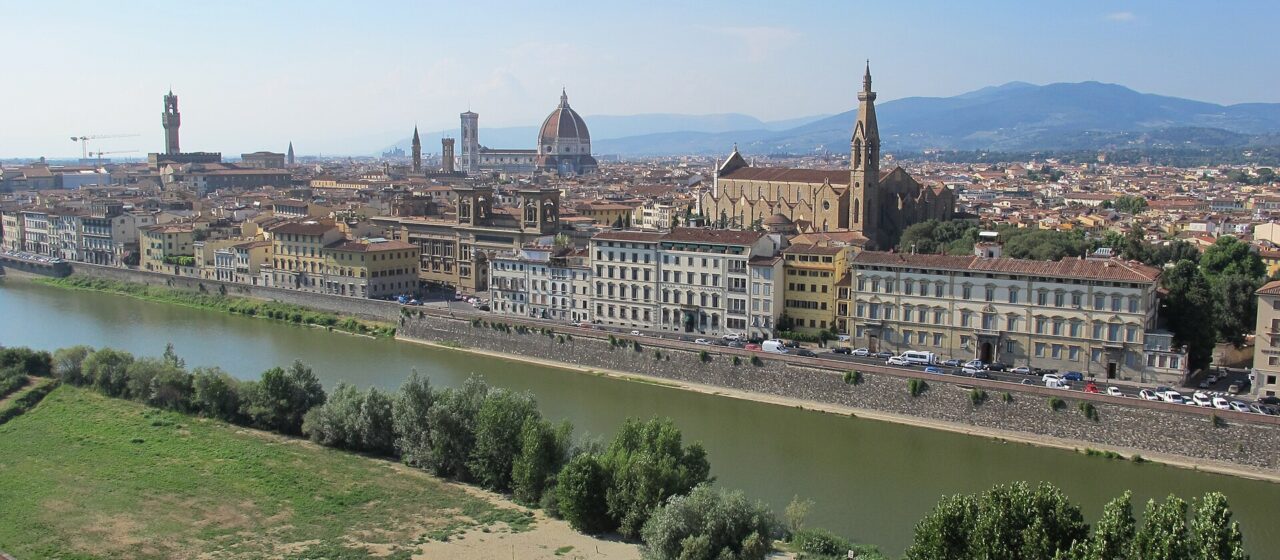 The Arno curves past Florence's riverfront buildings beneath Santa Croce, the cathedral dome and distant hills in clear daylight.
