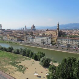 The Arno curves past Florence's riverfront buildings beneath Santa Croce, the cathedral dome and distant hills in clear daylight.
