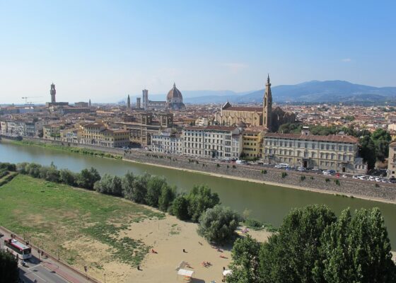The Arno curves past Florence's riverfront buildings beneath Santa Croce, the cathedral dome and distant hills in clear daylight.