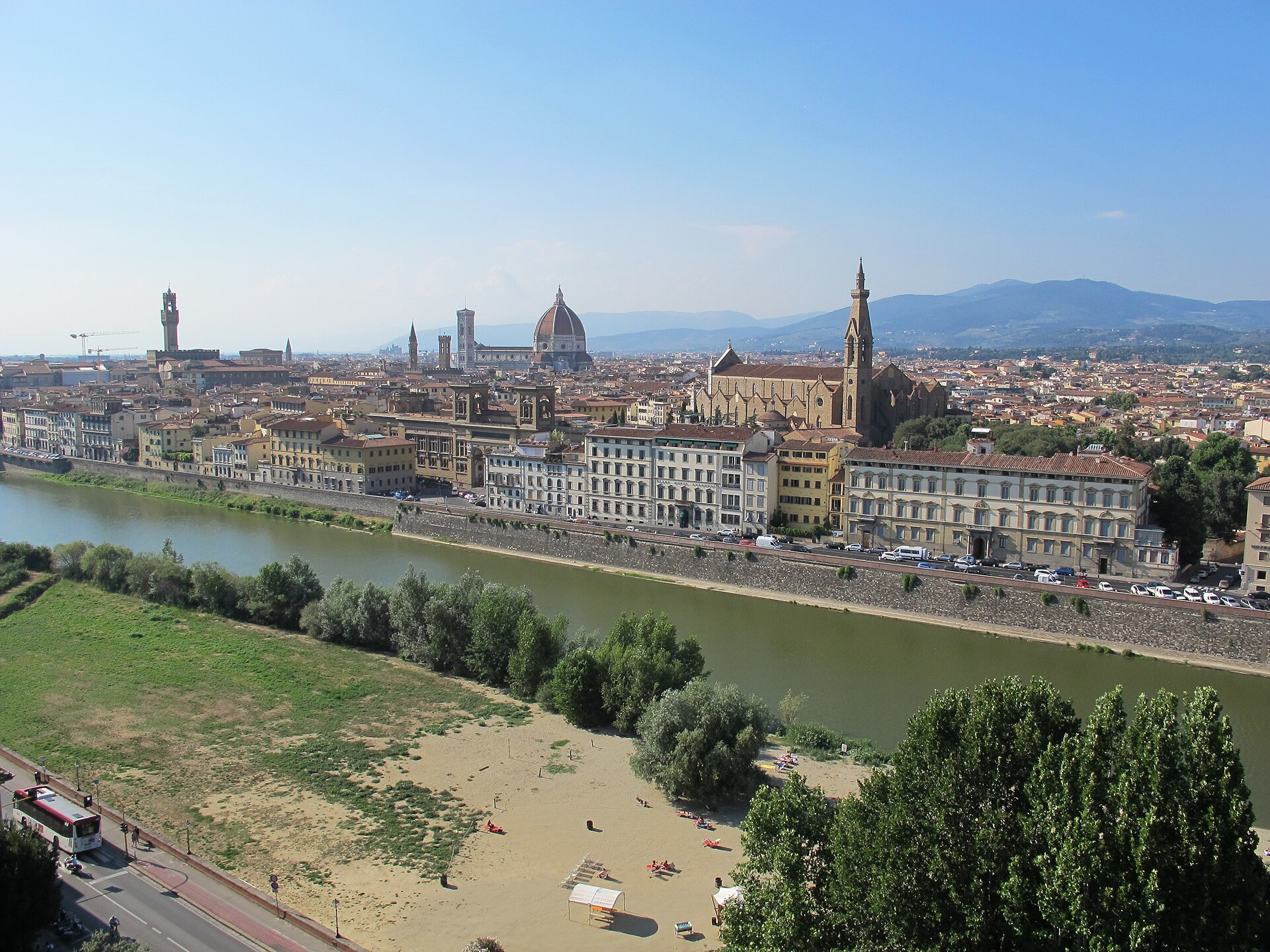 The Arno curves past Florence's riverfront buildings beneath Santa Croce, the cathedral dome and distant hills in clear daylight.