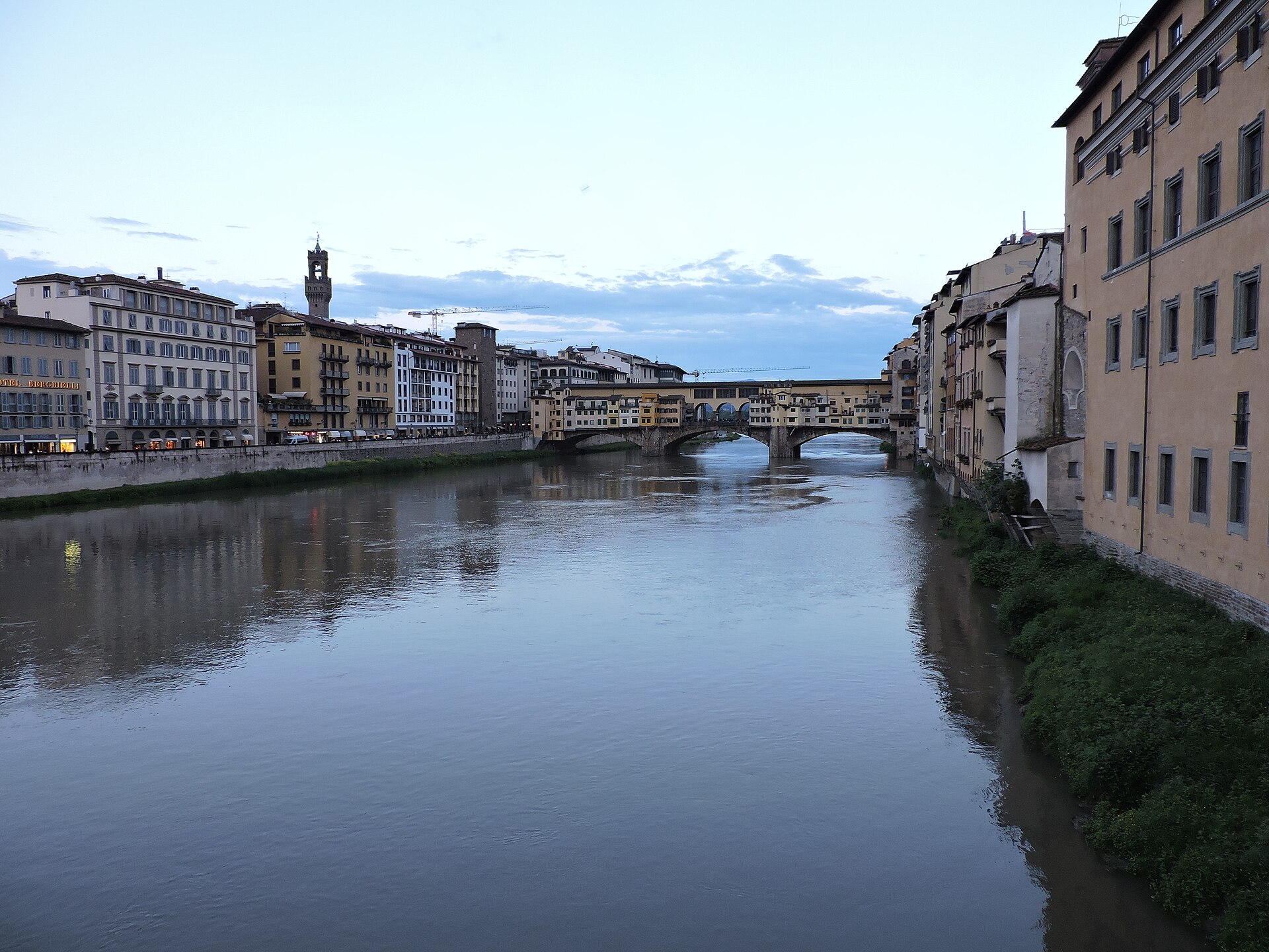 The Arno leads toward Ponte Vecchio in Florence at blue hour, with riverside buildings and Palazzo Vecchio’s tower framing the bridge.