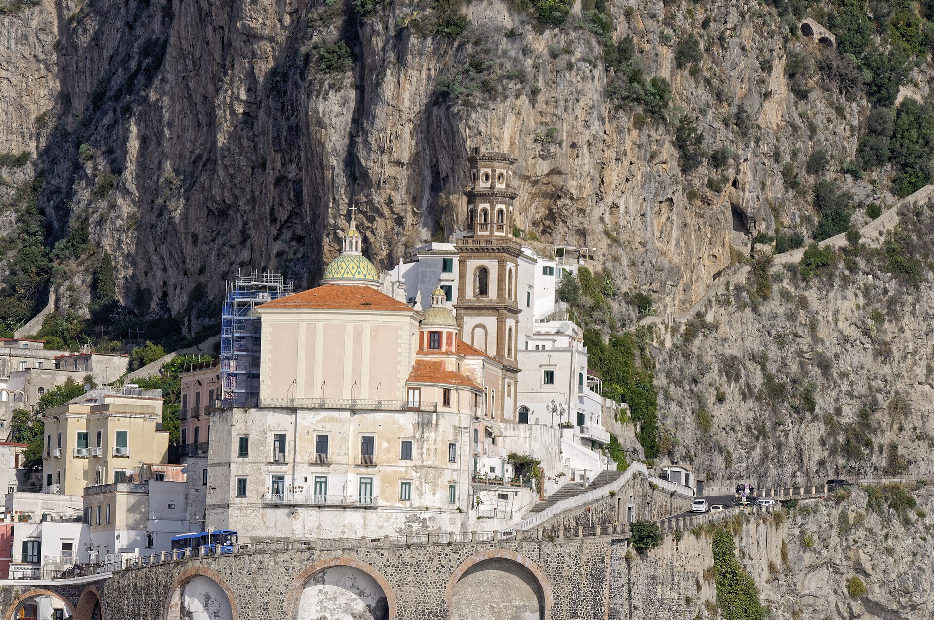 A church with a tiled dome and bell tower in Atrani, built against a sheer grey cliff above the arches of the coast road