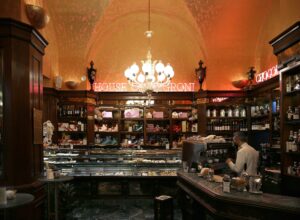 Barman behind the dark wood counter of Rivoire, a historic Florence cafe-bar, with shelves of bottles and chocolates under a vaulted ceiling and chandelier