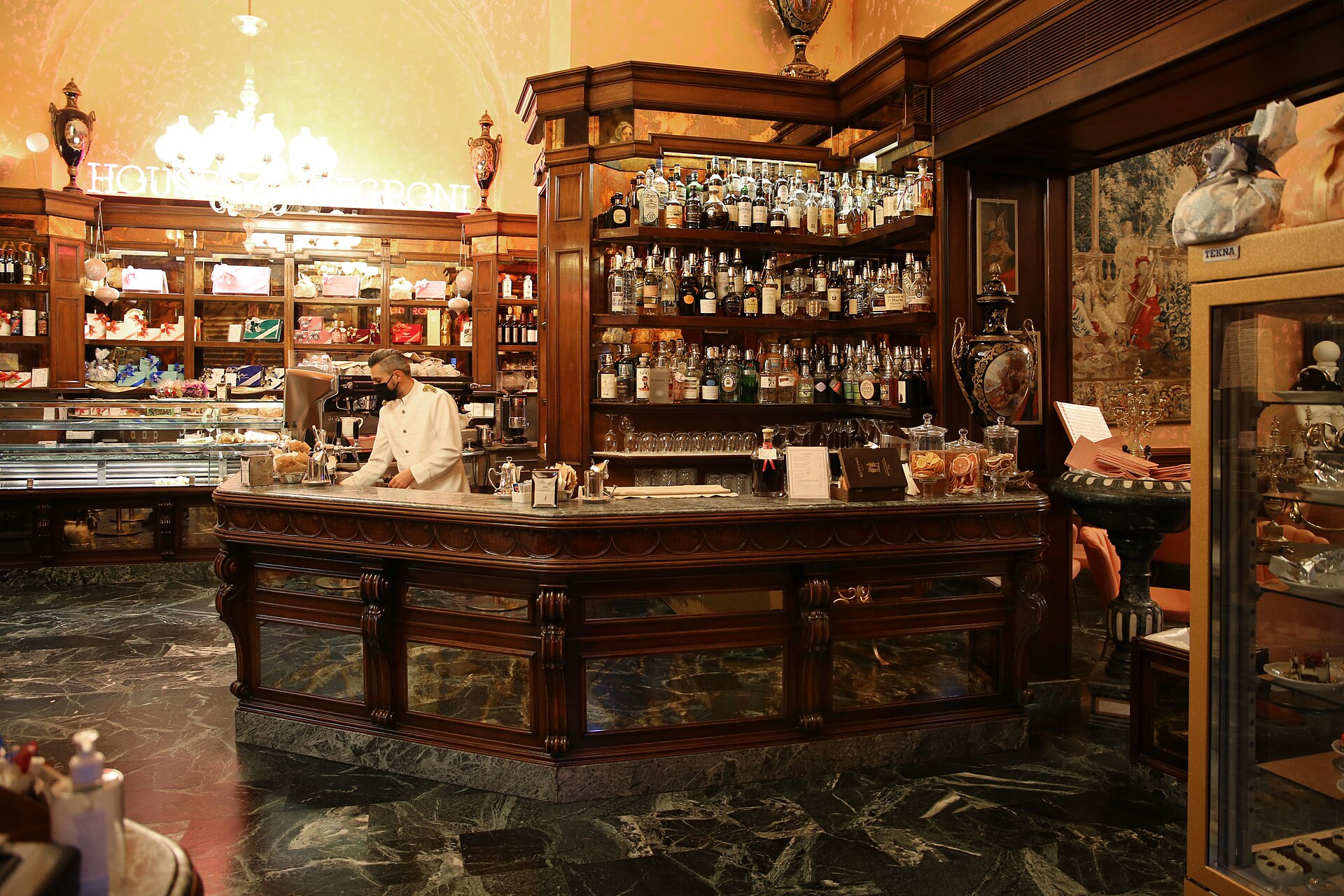 A bartender in a white jacket stands behind a curved wooden bar lined with spirits bottles at Rivoire in Florence, under a neon sign.