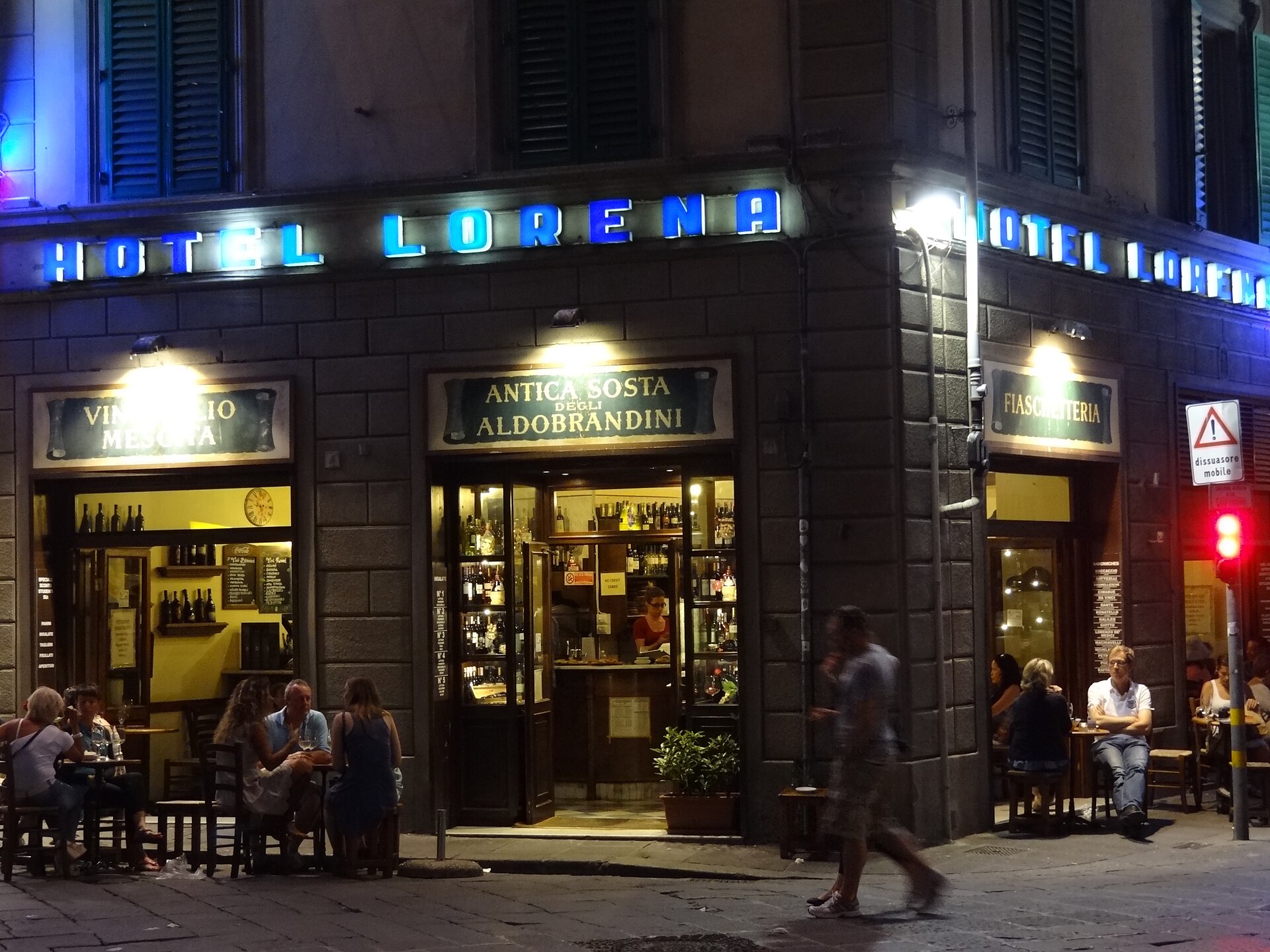 Blue Hotel Lorena lettering lights a rusticated Florence building above cafe windows and outdoor tables after dark.