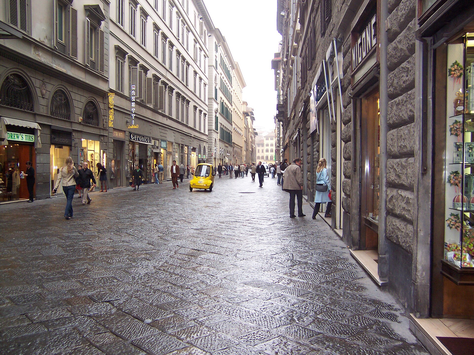 Broad gray stone slabs pave Via dei Calzaiuoli in Florence, with pedestrians walking between tall historic buildings.