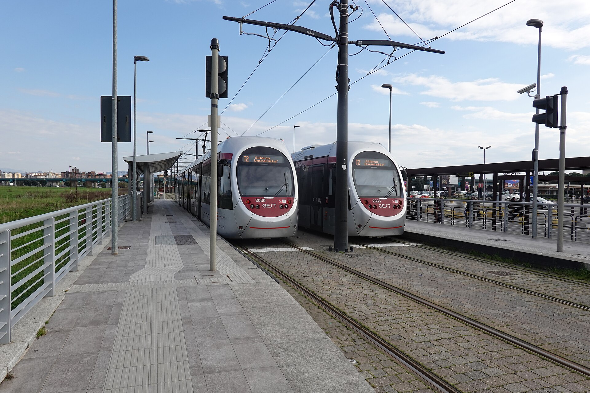 A broad paved platform leads to two trams at Florence Airport, with shelters and overhead wires beside the tracks.