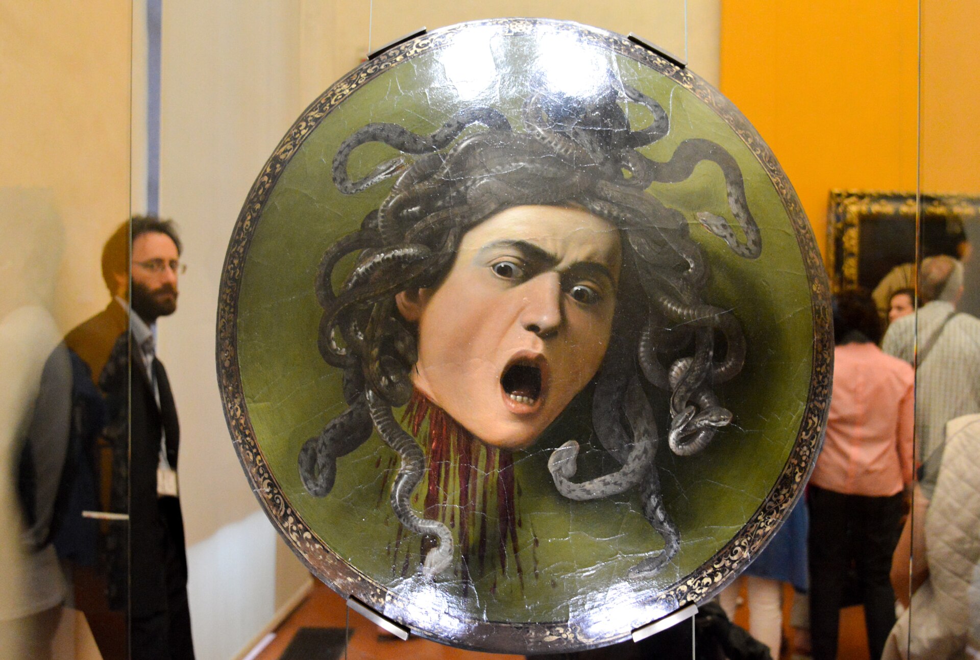 Caravaggio's snake-haired Medusa fills a round shield displayed behind glass at Florence's Uffizi Gallery, with visitors in the background.
