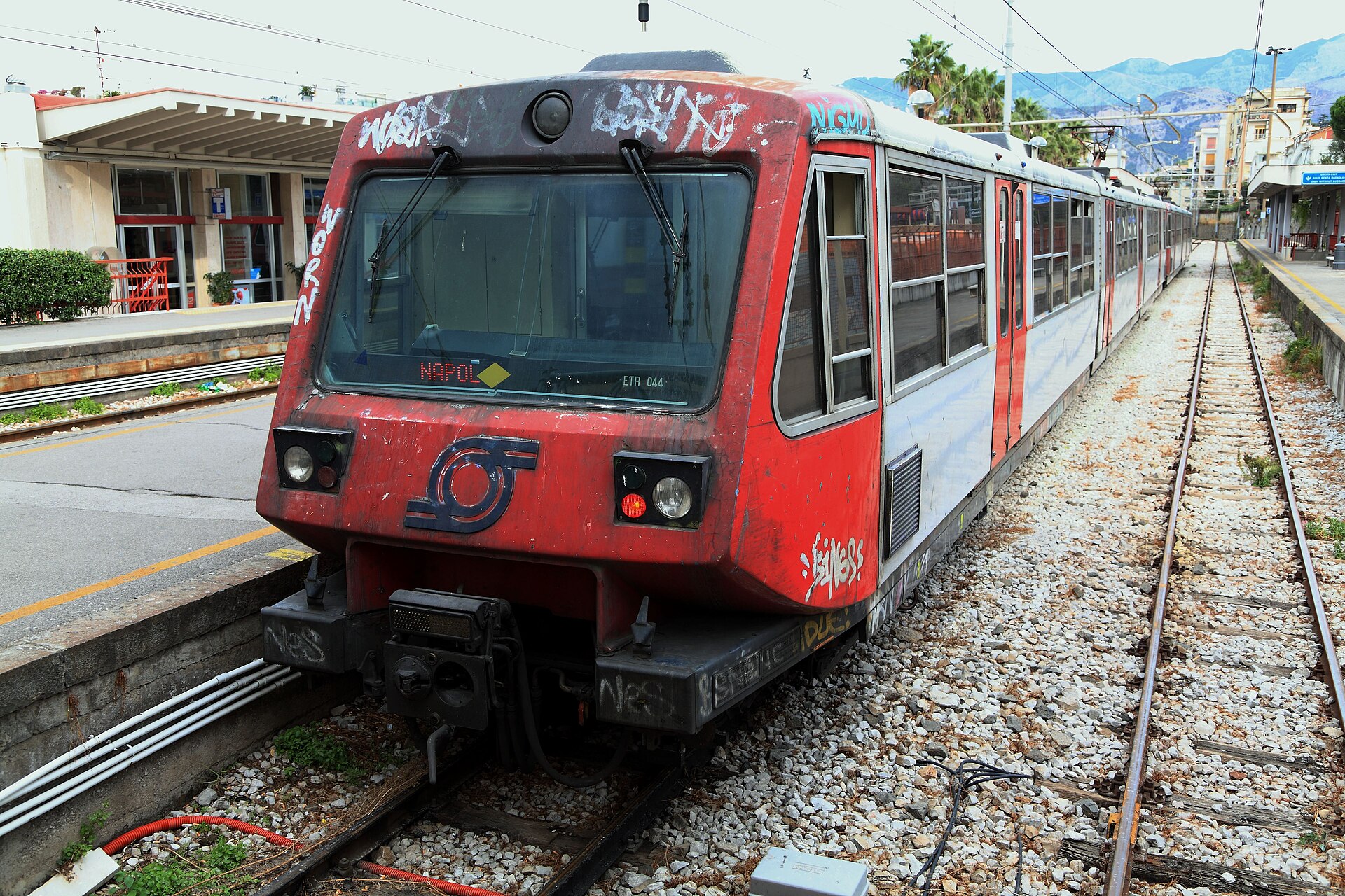 A red and white Circumvesuviana electric train, its front marked with graffiti, standing at the platform of Sorrento station with palm trees beyond