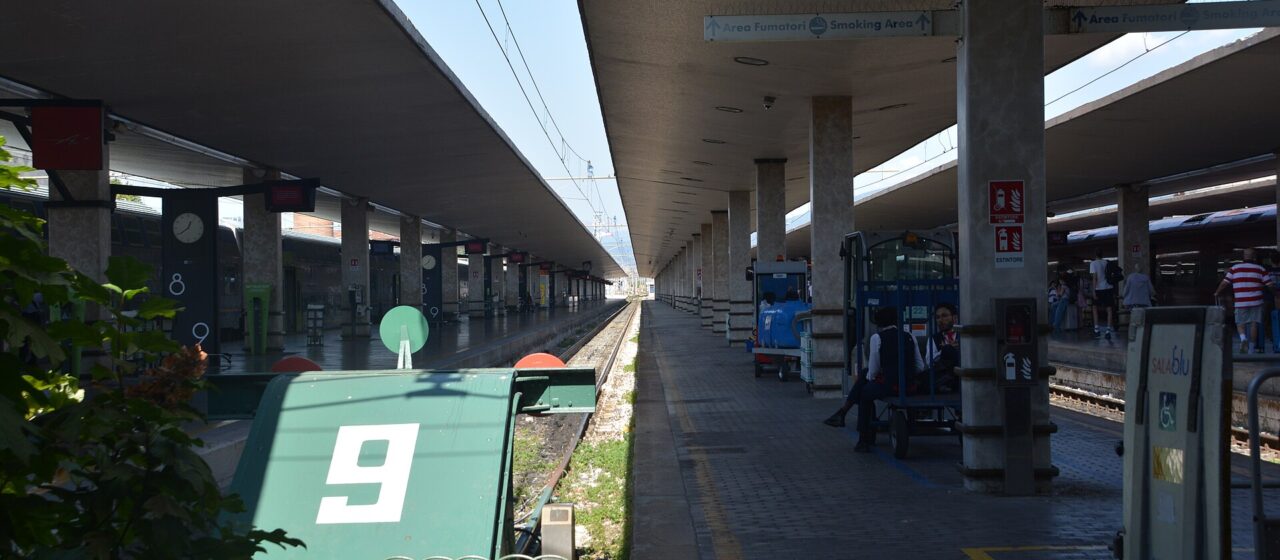 Concrete canopies shade Santa Maria Novella’s platforms in Florence, with track buffers and waiting passengers beside a train.