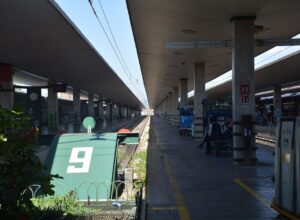 Concrete canopies shade Santa Maria Novella’s platforms in Florence, with track buffers and waiting passengers beside a train.